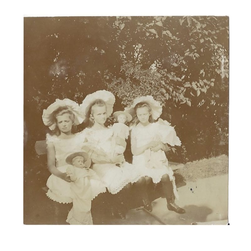 A photograph of Grand Duchesses Olga, Tatiana, & Maria holding dolls & seated on a bench in the gardens at Peterhof.